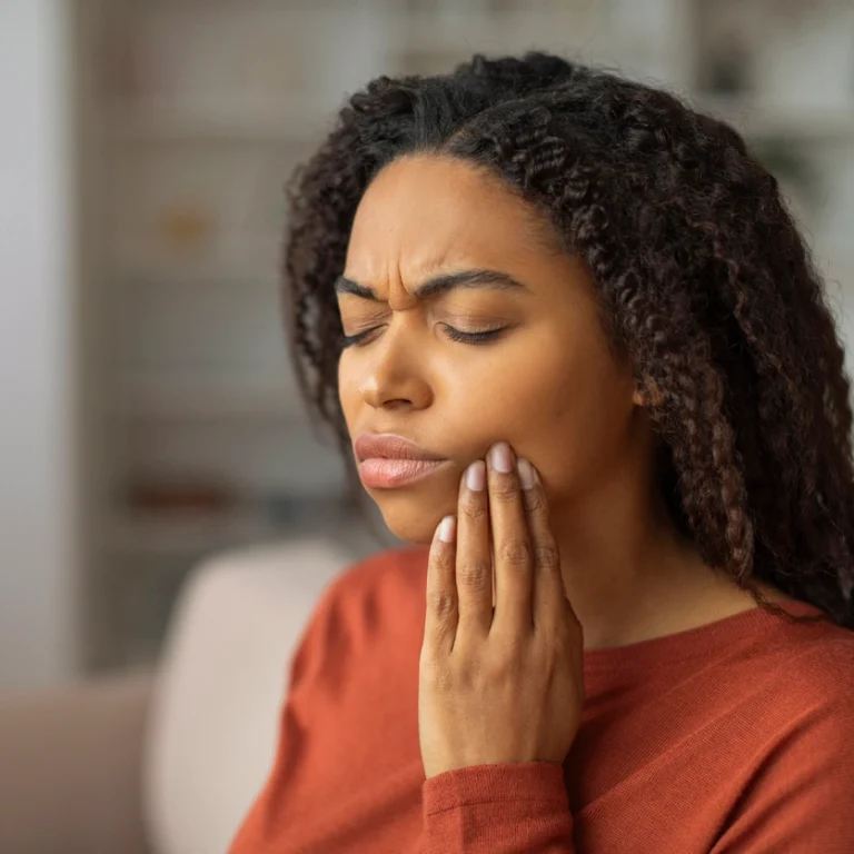 A woman at home holding her cheek in pain, visibly distressed, experiencing a dental emergency in a living room setting
